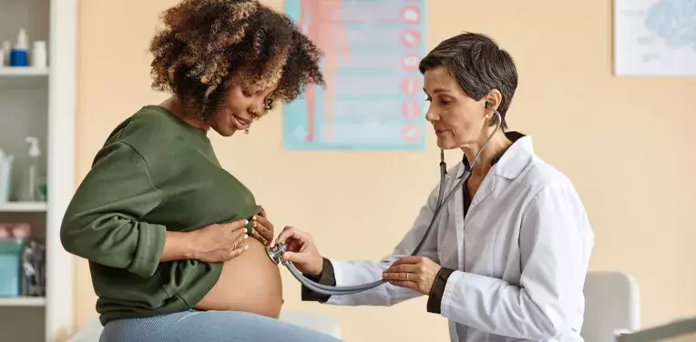 Side view of young pregnant woman looking at her belly while a doctor is checking baby with stethoscope in modern clinic