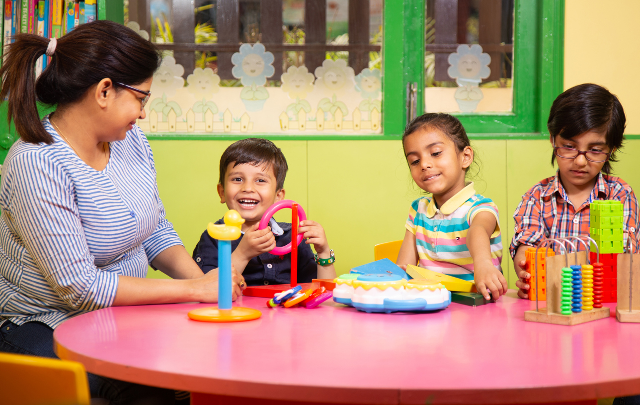 Three young children playing around a table, they are being supervised by a woman