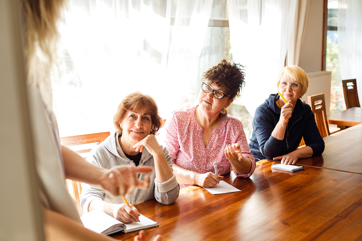 Three older women sat at a table taking notes and listening to a person speaking